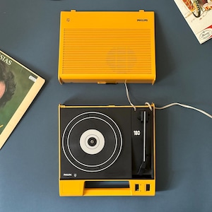 以下が含まれることがあります： 鮮やかな黄色いヴィンテージのPhilipsポータブルレコードプレーヤー。黒いターンテーブルと、それに合わせたスピーカーが特徴です。ターンテーブルには白と黒のレコードが載っています。スピーカーはターンテーブルの上にあります。背景にはレコードが見えます。