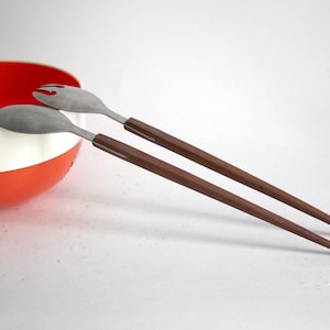 Puede incluir: Un juego de dos cucharas para ensalada con mangos de madera marrón y cabezas de metal plateado. Las cucharas están sobre una superficie blanca junto a un tazón rojo y blanco.