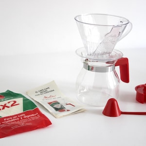 Puede incluir: Un juego de cafetera de vidrio transparente con asa, cuchara y tapa rojas. Incluye un cono de filtro y dos paquetes de filtros de café. Un paquete es verde y rojo con el texto "1x2".