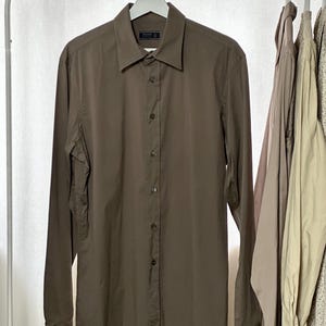 Pode incluir: Uma camisa marrom de manga comprida está pendurada em um cabide branco. A camisa tem uma gola clássica e uma fileira de botões na frente. O nome da marca é visível. Outras camisas são visíveis ao fundo.