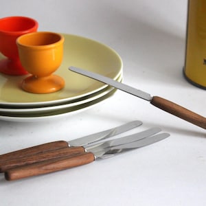 Puede incluir: Naturaleza muerta con tres cuchillos de mantequilla con mangos de madera y hojas de acero inoxidable. Dos hueveras, una naranja y otra amarilla, sobre una pila de platos verde claro. Una lata amarilla con texto en el fondo.
