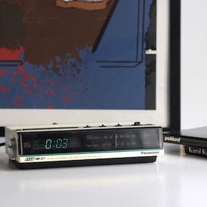 Op de afbeelding: Een vintage Panasonic AM/FM wekkerradio met een rechthoekige behuizing van gebroken wit en zwart plastic. De voorzijde bevat een digitaal groen LED-display dat 0:03 aangeeft, een analoge radiofrequentie-afstemknop en de tekst Easy Time Set en Panasonic.