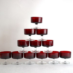 Puede incluir: Un juego de 12 copas de cristal transparente con tallo y cuenco rojo oscuro. Las copas están dispuestas en forma de pirámide.