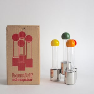 Puede incluir: Un juego de seis vasos de chupito de vidrio transparente con tapas de colores rojo, amarillo y verde. Los vasos están dispuestos en círculo y se encuentran sobre una base plateada. Los vasos están en su caja original, que es marrón con el texto "berndorff schnapsbar" impreso en rojo.