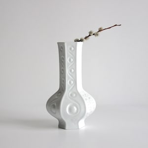 May include: A white ceramic vase with a unique shape and decorative embossed patterns. The vase has a tall, slender neck and a bulbous base. A sprig of pussy willow is placed in the vase. The vase is a decorative home decor item.