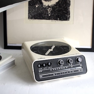 Puede incluir: Un radio despertador vintage blanco y negro con una esfera redonda y una pantalla digital. La radio tiene un dial con bandas AM, MW y UKW. La radio está etiquetada como "Quartz Electronic CR-15".