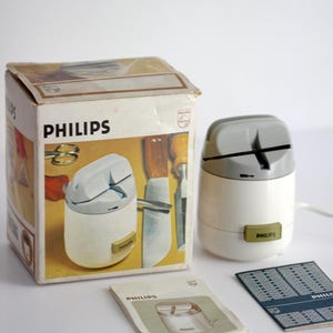Könnte beinhalten: Ein Vintage-Messerschärfer von Philips, weiß und grau, mit Originalverpackung und Papieren. Die Box zeigt eine Abbildung des Schärfers und der Werkzeuge. Der Schärfer hat ein kleines grünes Etikett mit dem Philips-Logo.