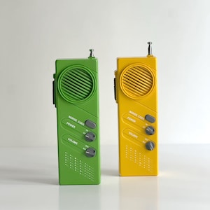 Puede incluir: Dos dispositivos walkie-talkie rectangulares de mano en plástico verde brillante y amarillo. Cada unidad cuenta con una rejilla de altavoz circular, una pequeña antena de metal, un botón de código Morse, un interruptor y un dial de volumen.