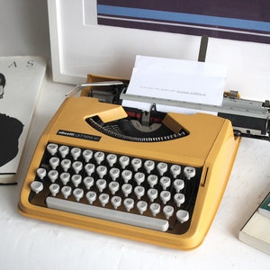 May include: A yellow vintage Olivetti Lettera 32 typewriter with a sheet of paper in the carriage. The typewriter is on a white surface with two books in the background.