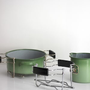 Puede incluir: Una olla de metal esmaltado verde y una jarra a juego, ambas con interior gris y un soporte de alambre cromado extraíble con agarres laterales de plástico negro. La jarra muestra una pequeña etiqueta rectangular con el texto SCHIRR.
