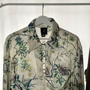 Puede incluir: Camisa abotonada de color blanquecino con estampado floral y de pájaros. La camisa tiene cuello y mangas largas. El diseño incluye motivos florales y de pájaros azules, verdes y morados. La camisa está colgada en una percha blanca.