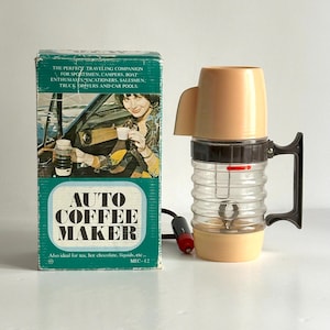 以下が含まれることがあります： ベージュの蓋と透明なリブ付きカラフが付いた、ヴィンテージの自動コーヒーメーカー。黒いハンドルと電源コードが見えます。「AUTO COFFEE MAKER」の文字と、車に乗っている人のイラストが描かれた箱。