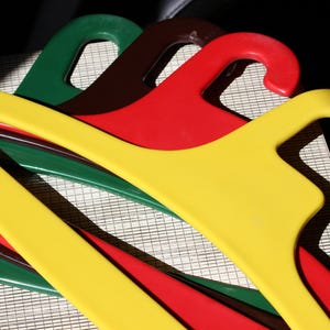 May include: A close-up of several colourful plastic clothes hangers. The hangers are in shades of yellow, red, green, and brown. The hangers are overlapping each other. The background is a light-coloured surface with a grid pattern.