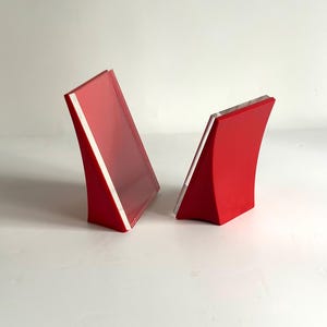 Puede incluir: Dos sujetalibros rojos curvados con un borde blanco. Los sujetalibros tienen un diseño moderno y minimalista y están colocados sobre un fondo blanco. Están hechos de un material sólido y tienen un acabado brillante.