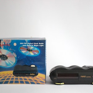Könnte beinhalten: Ein schwarzes AM/FM Digital-Radio mit eingebauter Nachtbeleuchtung. Das Radio steht auf einer weißen Oberfläche und hat ein rotes digitales Display. Die Verpackung des Radios ist blau und zeigt ein Bild einer CD. Auf der Verpackung steht auch "FIRST AUSTRIA" und "New Wave".