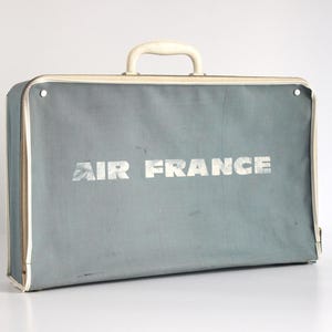 Peut inclure: Valise Air France vintage de couleur bleu-gris. Elle présente une poignée et des bordures crème, avec le logo "AIR FRANCE" en lettres blanches. La fermeture éclair est dorée.