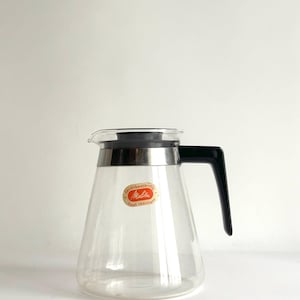 Puede incluir: Una jarra de café de cristal transparente con tapa y asa de plástico negro. La jarra presenta una banda metálica de color plateado alrededor del borde superior y una pegatina ovalada de color naranja con el texto Melitta, feuerbeständig y heat resistant.