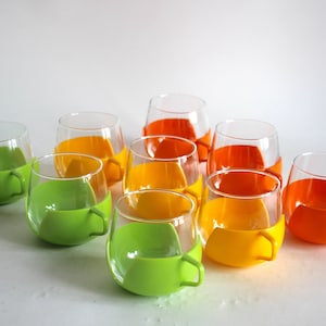 Può includere: Set di otto tazze di vetro con porta tazze in plastica colorata. Le tazze sono in vetro trasparente e i porta tazze sono verdi, gialli e arancioni.