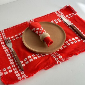 Puede incluir: Un mantel individual rojo con bordado floral blanco, un plato de madera, una servilleta roja con un anillo de madera, un tenedor y un cuchillo con mangos de madera. El mantel individual tiene un borde deshilachado. Vajilla.