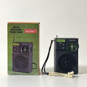 May include: A retro black AM/FM solar-powered pocket radio and its original box. The radio has a telescopic aerial, a speaker grille, and a tuning dial. The box is green and reads "AM/FM SOLID STATE POCKET RADIO".