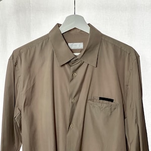 Pode incluir: Camisa bege com botões, gola clássica e mangas compridas. A camisa tem um bolso no peito com uma etiqueta preta e uma etiqueta pequena perto da gola. A camisa está pendurada em um cabide branco.