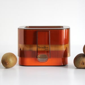 Può includere: Un elettrodomestico da cucina arancione e marrone in stile retrò con la scritta "Terraillon 4000". Diversi kiwi sono disposti attorno all'elettrodomestico. L'elettrodomestico ha una forma rettangolare con una parte superiore argentata.
