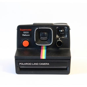 Puede incluir: Una c&aacute;mara Polaroid Land negra con un bot&oacute;n rojo y una franja arco&iacute;ris. La c&aacute;mara tiene una etiqueta Supercolor 1000 Deluxe.