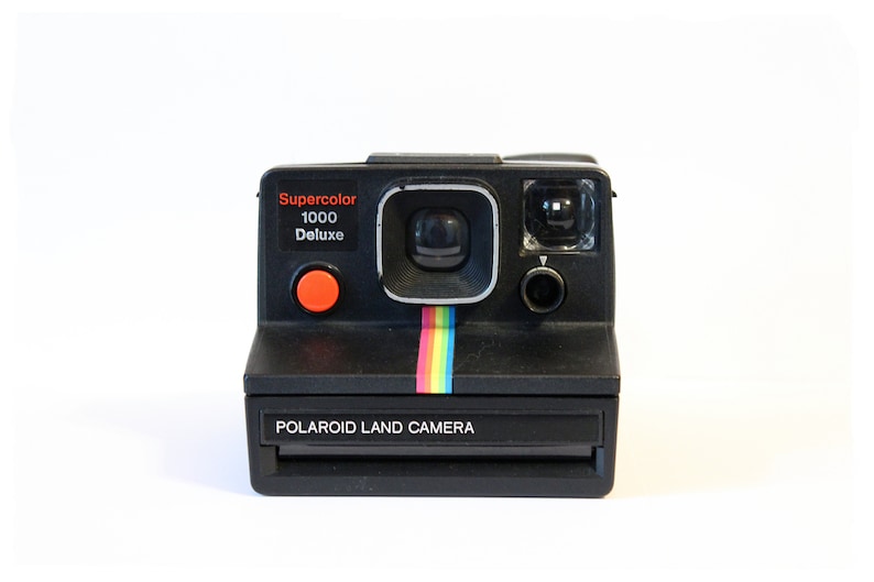 Puede incluir: Una c&aacute;mara Polaroid Land negra con un bot&oacute;n rojo y una franja arco&iacute;ris. La c&aacute;mara tiene una etiqueta Supercolor 1000 Deluxe.
