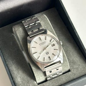 Könnte beinhalten: Eine silberne Citizen Automatic Armbanduhr mit silbernem Armband, präsentiert in einer schwarzen Box mit grauem Samtinnenraum. Das Zifferblatt der Uhr ist silberfarben mit schwarzen Zeigern und Markierungen, und das Citizen-Logo ist sichtbar.