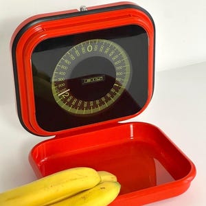 Puede incluir: Una báscula de cocina roja vintage con una esfera negra y un dial circular que muestra las mediciones de peso. La báscula tiene una bandeja roja a juego y está acompañada de un racimo de plátanos amarillos. El dial de la báscula lee hasta 1000 gramos.
