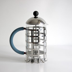 Puede incluir: Una cafetera de prensa francesa de acero inoxidable con un mango azul y una jarra de vidrio transparente. La jarra tiene un filtro de malla de acero inoxidable.