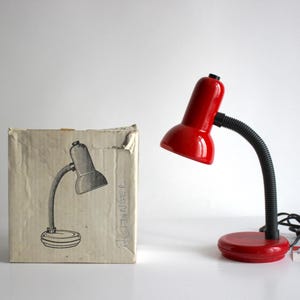 Puede incluir: Una lámpara de escritorio roja con cuello flexible y cable negro. La lámpara está en su caja original, que es blanca con texto negro.