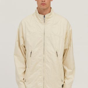 May include: A light beige jacket with a high collar and a full-length zipper. The jacket has two side pockets with zippers and a small logo near the bottom. The model is wearing a dark navy blue pair of pants.