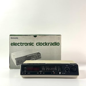 May include: A vintage Philips electronic clockradio with a black and cream color scheme. The clock radio displays the time as 10:32. The original box is behind the clock radio and has the words "electronic clockradio" printed on it.