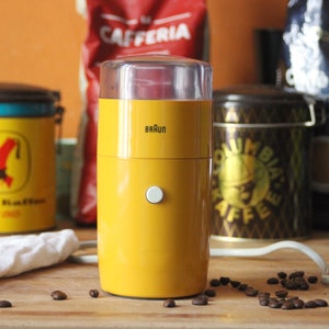 Pode incluir: Um moedor de café elétrico Braun amarelo com tampa transparente e botão branco. Grãos de café espalhados em uma superfície de madeira. Latas e sacos de café ao fundo.