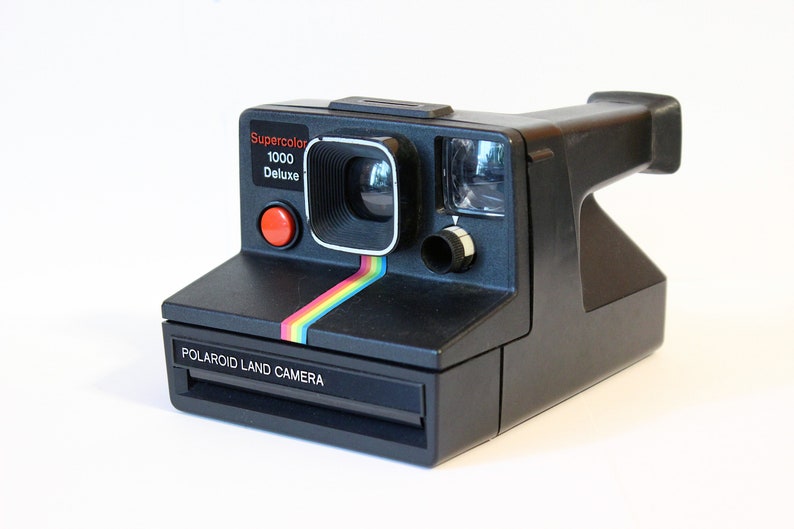 Puede incluir: Una c&aacute;mara Polaroid Land negra con una franja arco&iacute;ris en la parte delantera. La c&aacute;mara tiene un bot&oacute;n rojo y el texto "Supercolor 1000 Deluxe" en la parte delantera.
