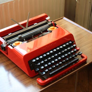 Peut inclure: Une machine à écrire vintage rouge vif avec des touches noires et le nom "Valentine" sur le devant. La machine à écrire est posée sur une table en bois, mettant en valeur son design rétro. Les touches sont noires et les accents sont orange.