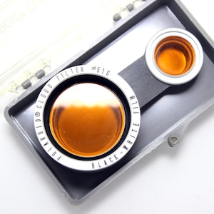 Puede incluir: Primer plano de una caja de filtros Polaroid con dos filtros en su interior. Los filtros están etiquetados como "Polaroid 01 Cloud" y "Black-White Film".