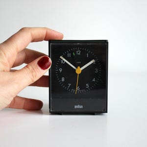 Può includere: Un orologio da tavolo Braun nero con quadrante bianco e lancette gialle. L'orologio ha un design semplice ed è impostato alle 10:50.