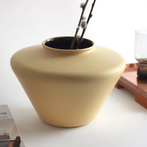 Puede incluir: Un jarrón de cerámica de color amarillo pálido con una boca ancha y una base redondeada. El jarrón está sobre una superficie blanca con un libro y una bandeja con vasos.
