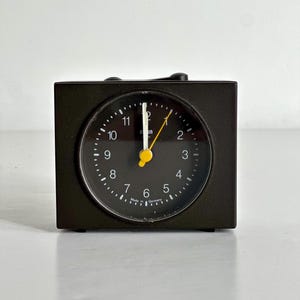 Puede incluir: Un reloj despertador analógico negro y rectangular con una esfera circular. La esfera tiene números blancos y un segundero amarillo. La marca "Braun" es visible. El reloj está sobre una superficie blanca.