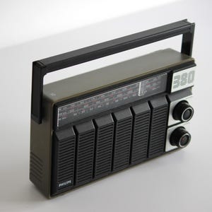 Könnte beinhalten: Ein Vintage-Radio Philips 380 AM/FM mit schwarzem Griff und einem grün-schwarzen Gehäuse. Das Radio hat ein Zifferblatt mit Zahlen und einen Lautsprecher mit einem Gitter.