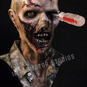 May include: A horrifying zombie bust with a screwdriver through its eye. The zombie has a pale complexion, open wounds, and a decaying appearance. The bust is mounted on a round base.