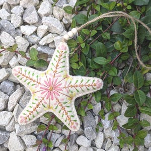 May include: A cream-coloured felt star ornament featuring intricate hand-stitched embroidery in shades of pink, yellow, and green. The star is suspended by a jute twine string adorned with three small, round, light-coloured wooden beads.