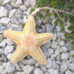 May include: A cream-coloured felt starfish ornament featuring intricate hand-stitched embroidery in shades of pink, orange, and green. The starfish is suspended by a jute twine string adorned with three small, round, light-coloured wooden beads.
