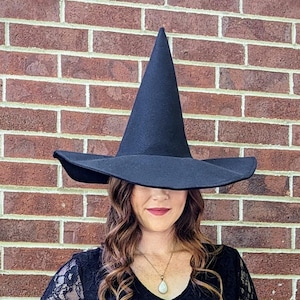 Könnte beinhalten: Ein schwarzer Hexenhut mit breiter Krempe und hoher, spitzer Krone. Der Hut wird von einer Person getragen, die ein schwarzes Spitzenkleid trägt. Der Hintergrund ist eine Backsteinmauer.