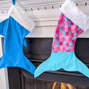 May include: Two Christmas stockings hanging on a fireplace mantel. One stocking is blue and shaped like a shark fin, the other is pink and turquoise and shaped like a mermaid tail.