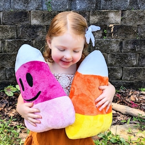 Könnte beinhalten: Ein Kind hält zwei Plüsch-Zuckermais-Kissen. Eines ist rosa mit einem lächelnden Gesicht, das andere ist orange und gelb. Beide haben weiße Spitzen. Die Kissen sind weich und farbenfroh, ideal für Halloween oder ein verspieltes Zimmer.