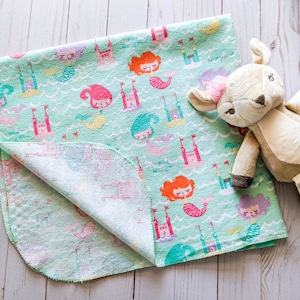 May include: A soft, green baby blanket with a mermaid and castle print. The blanket is folded in half and a plush fawn toy is lying on top of it.