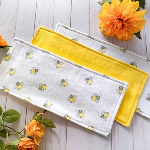 May include: Three fabric burp cloths with a white background and a yellow bee pattern. The top two cloths are yellow and the bottom cloth is white.